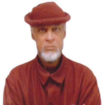 Joseph "Jo-Jo" Bowen in red prison uniform with red cardigan and hat, white beard, looking at camera, unsmiling