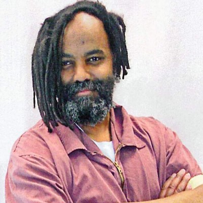 Mumia Abu-Jamal looking at camera, smiling with some white in his beard, with arms crossed