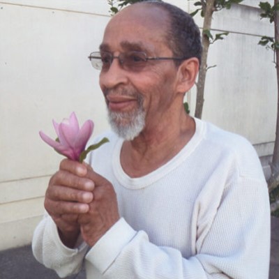 Veronza Bowers holding a flower outside, smiling, white beard, not looking at camera