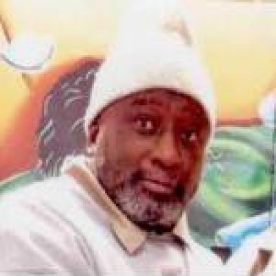 Kobo Bomani Sababu in a tan collared shirt with a white top over it and a white hat, with some white in his beard, looking a camera in front of colorful background